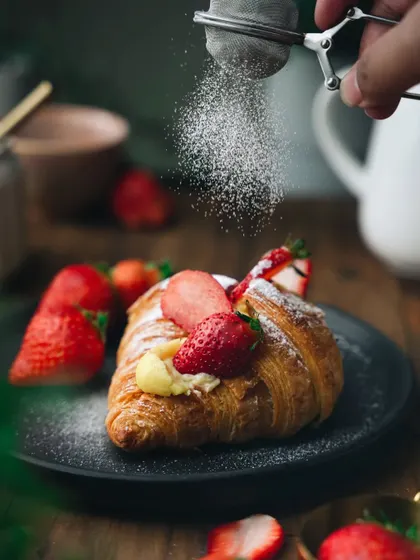 A moody, dramatic shot of a strawberry custard croissant being dusted with powdered sugar. The dark background makes the flaky pastry and bright red strawberries stand out beautifully.