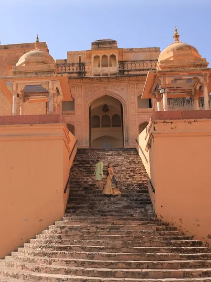 A wide-angle shot of a couple ascending the grand stone staircase of a historic Indian palace. This image captures the scale and majesty of destination pre-wedding locations.