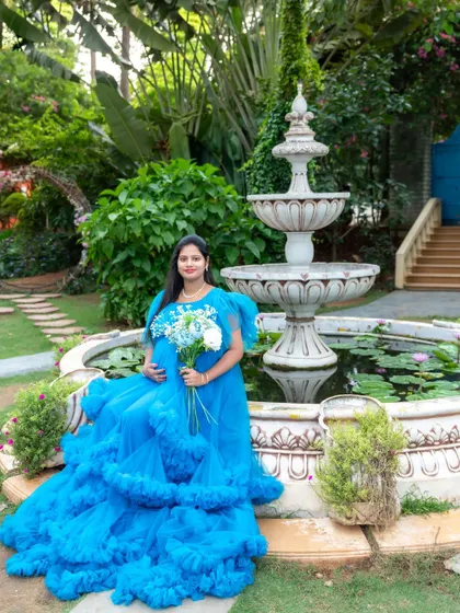 A beautiful portrait of a mom-to-be in a blue ruffled gown, sitting by a garden fountain and holding a bouquet of flowers.