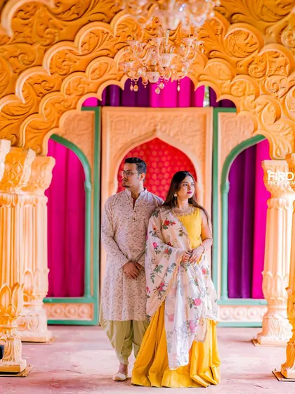 A regal and poised portrait set against a grand, colorful archway. The couple's traditional yellow and white outfits look stunning in this architectural setting.