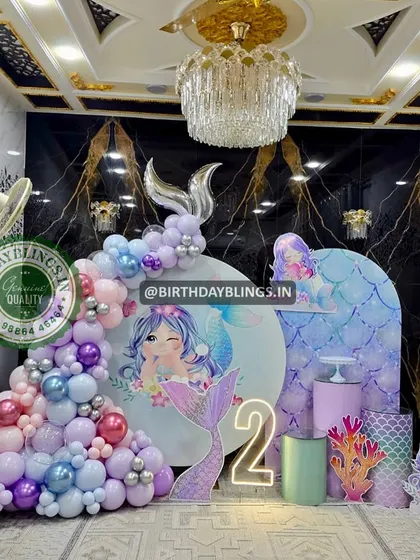 A glamorous mermaid-themed second birthday party set against a marble wall. The decor features iridescent and pastel balloons, a neon number '2', and shimmering mermaid tail cutouts.