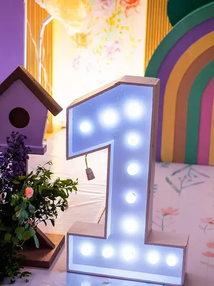 A light-up number '1' prop next to a colorful rainbow and birdhouse details. I use illuminated props to add a warm and celebratory glow to the party decor.