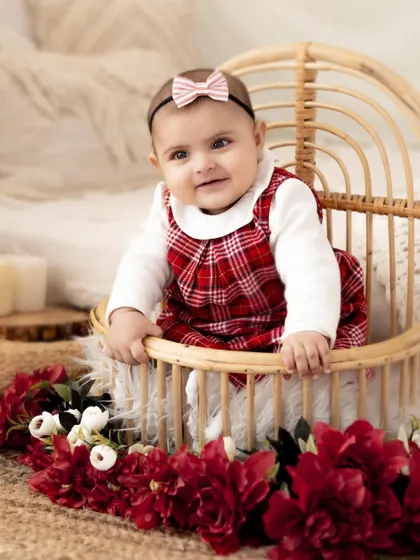 A sweet baby girl in a festive red plaid dress, sitting in a rustic wicker chair. This setup is perfect for a sitter session or a holiday-themed shoot.