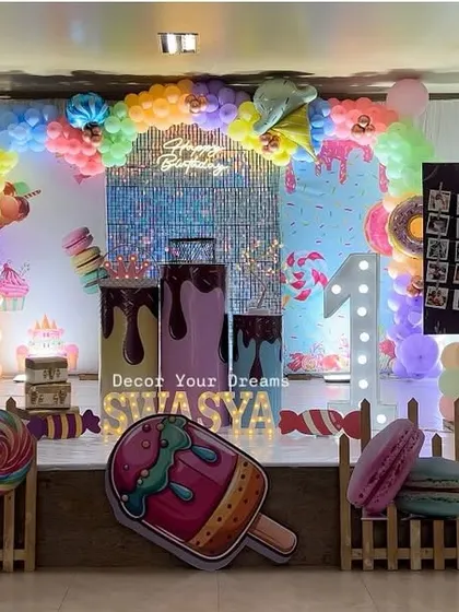 A sweet candy-themed birthday celebration. This elaborate setup includes a shimmer wall, large cutouts of ice cream and donuts, a colorful balloon arrangement, and a picket fence.