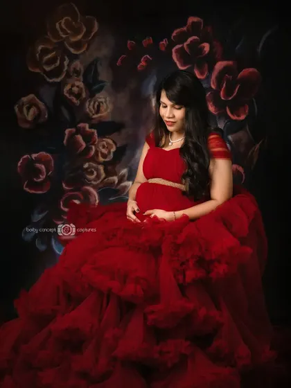 A seated pose from the red gown series, showing the mom-to-be cradling her bump. The voluminous red tulle spreads around her against the floral backdrop, creating a beautiful, painterly effect.