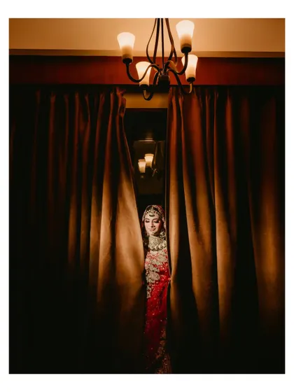 An artistic shot of the bride peeking from behind a curtain. The moody lighting and creative composition create a sense of mystery and romance.
