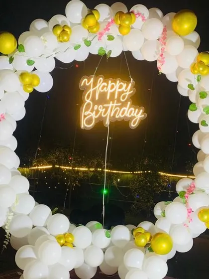 An outdoor birthday photo booth with a circular balloon frame in white and gold. The "Happy Birthday" neon sign adds a warm glow to the evening celebration.