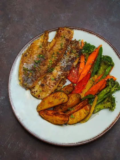 A healthy grilled fish platter, served with a side of roasted potatoes and mixed vegetables on a dark, rustic plate.