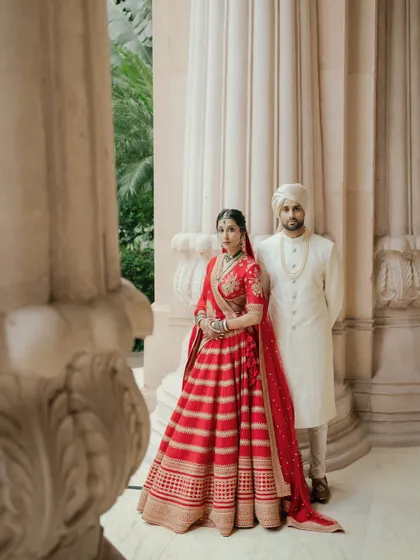 A classic portrait of Avin and Nikki at The Leela Palace, Bangalore. Their coordinated red and white outfits stand out beautifully against the palace's stately pillars.