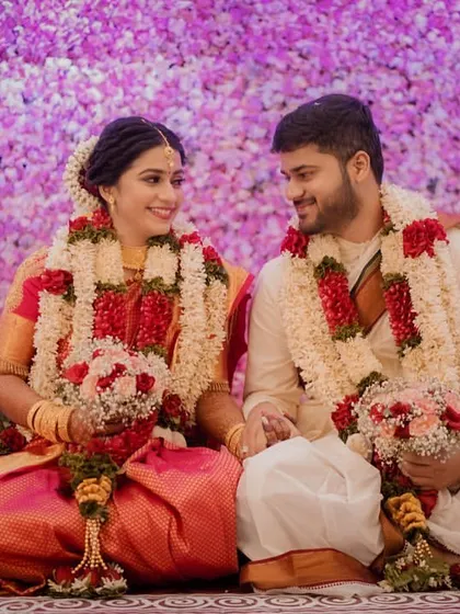 A beautiful portrait of the couple seated before a wall of purple flowers, holding bouquets and looking at each other with love.