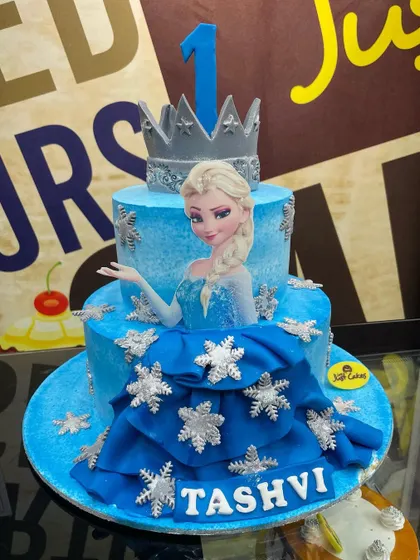 Let it go with this magical 4kg Frozen theme cake. This two-tiered creation features a fondant dress drape, an edible Elsa image, and sparkling snowflake details, perfect for a princess's party.