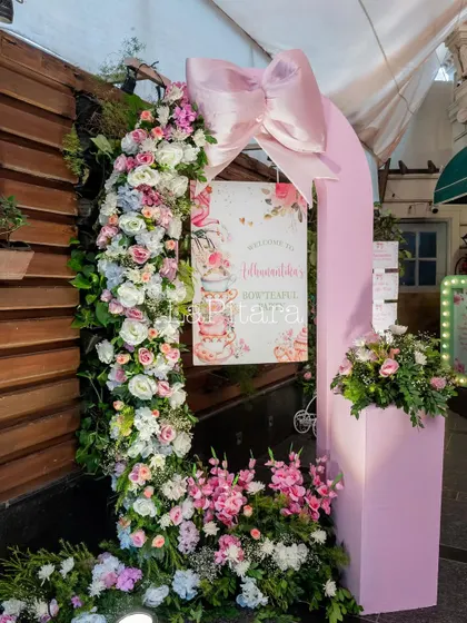The entrance arch for the bow-themed party, featuring a cascade of fresh flowers and a hanging welcome sign.