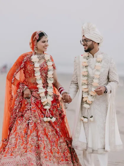 A classic couple portrait on the beach, holding hands and sharing a smile, with the bride's orange lehenga adding a pop of color against the serene backdrop.
