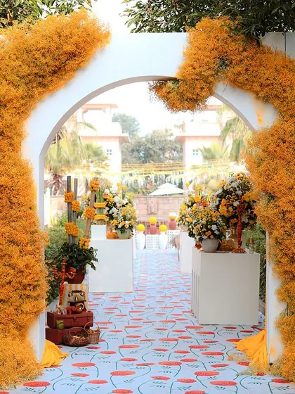 The entrance to the Haldi ceremony, featuring a white arch wrapped in fluffy, yellow-orange florals, leading onto a custom-printed poppy-patterned carpet.