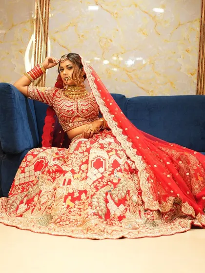 A full-shot of a bride on a couch, letting the magnificent red doli-themed lehenga spread out. The skirt depicts a traditional wedding procession, making it a unique and story-telling piece.