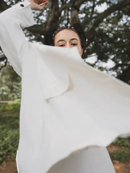 A shot from the 'SOLARA' editorial, focusing on movement and light. The flowing white shirt and gold-flecked makeup create an ethereal, free-spirited look against the natural backdrop of the trees.