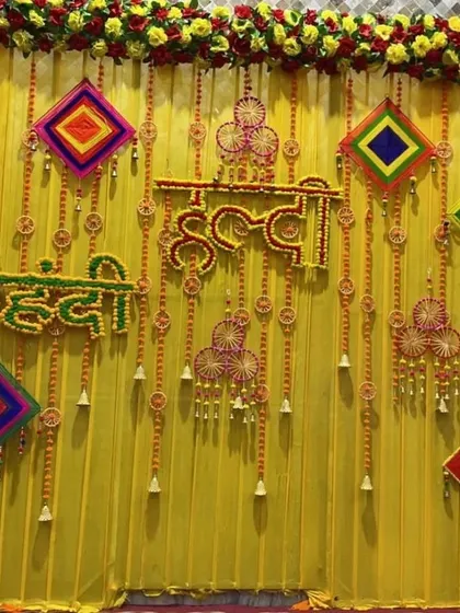 A combined 'Haldi Mehendi' backdrop in bright yellow, featuring floral signs for both functions and decorated with colorful hanging kites.