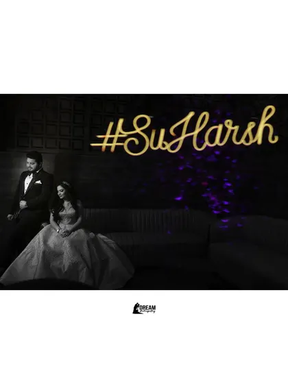 A black and white shot of Sunny and Harshna seated on a couch with their wedding hashtag #SuHarsh lit up behind them.