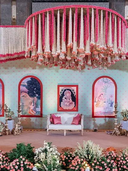 A grand and traditional stage for a South Indian half-saree function. The decor features massive floral chandeliers made of jasmine and pink flowers, with a custom backdrop depicting the stages of a woman's life.