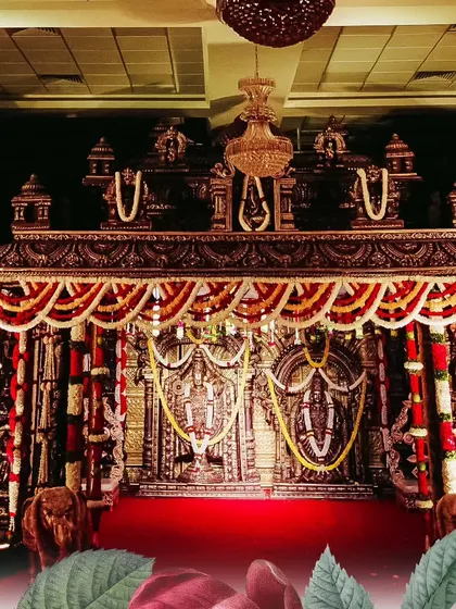 A full view of a traditional temple-themed wedding stage under elegant chandeliers. The intricate wooden-finish structure and floral arrangements create an atmosphere of timeless grandeur.