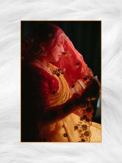 An artistic, moody portrait of a bride in a yellow and red saree, her face partially veiled, illuminated by soft, dramatic light.