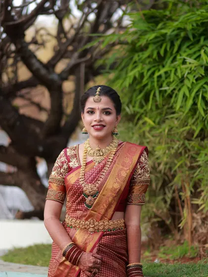 A full-length outdoor portrait. The bride's makeup and traditional attire look stunning against the natural green background.