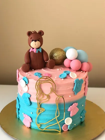 A "He or She" baby shower cake with a teddy bear topper and cute baby clothes details.