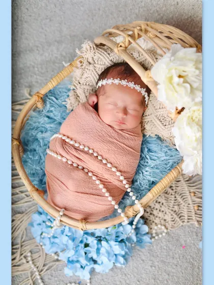 A newborn girl wrapped in a peach swaddle with pearls, sleeping peacefully in a round cane bassinet. The soft blue and white flowers provide a gentle, dreamy backdrop.