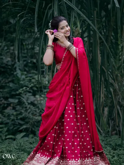 A beautiful portrait of the bride-to-be in a vibrant red lehenga, placing a flower in her hair amidst lush greenery.