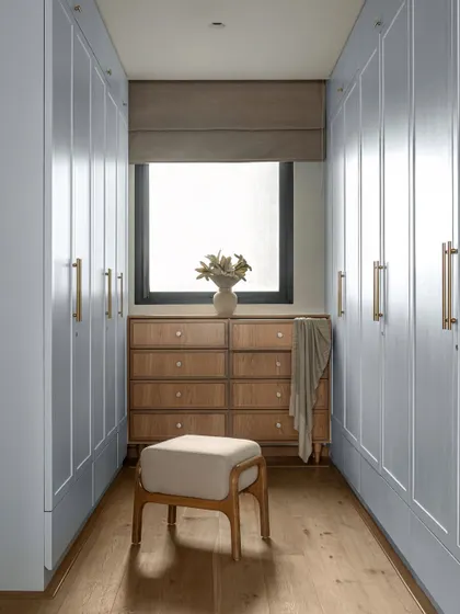 The walk-in closet in the KAIA master bedroom was designed to feel calm and functional. His-and-her wardrobes are finished in a soft blue that echoes the bedroom's highlights, while a custom wooden dresser sits under the window, bringing in natural light and a sense of grounded luxury.