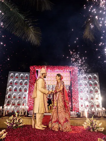 A moment of pure magic. The couple stands on a stage showered in flower petals, with fireworks lighting up the night sky behind them. This is the kind of grand, unforgettable varmala moment I love to create.