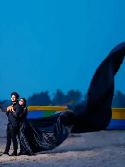 A dramatic beach pre-wedding shoot at dusk. The couple is coordinated in all black, with her long-trail gown catching the wind for a powerful and artistic shot.