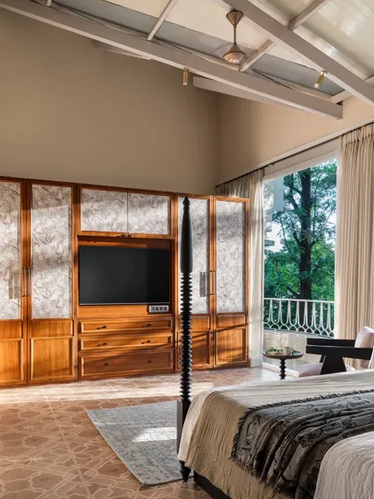 This spacious bedroom in the Panchgani villa features a high, beamed ceiling and a custom wooden media unit that conceals the TV. The large window opens up to a balcony with a beautiful green view.
