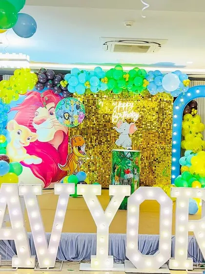 A fun jungle-themed first birthday stage featuring characters from The Lion King and Masha and the Bear. The setup includes a gold shimmer wall and large marquee letters spelling "AVYOR".