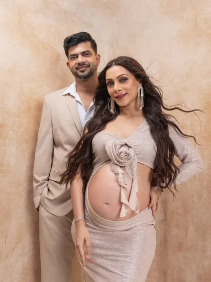 This is a chic and modern maternity look. The couple poses against a textured beige backdrop, with the mother-to-be showcasing her bump in a stylish, fitted two-piece outfit.
