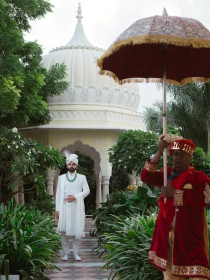 The groom makes a royal entrance, escorted under a traditional umbrella through the lush gardens of a palace hotel.