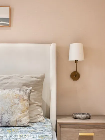 A close-up of the bedside shows the simple elegance of the design, with a classic wall-mounted lamp and a clean-lined nightstand. The textured wallpaper adds a layer of warmth.