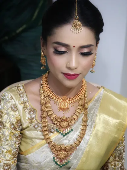 An elegant portrait of a South Indian bride. Her makeup is flawless, with soft, defined eyes and a classic red lip that complements her off-white and gold Kanchipuram saree and layered antique jewellery.