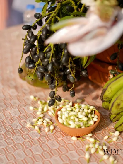 A close-up of a traditional fruit and flower arrangement, featuring bananas, jamun, and a bowl of fresh jasmine buds.