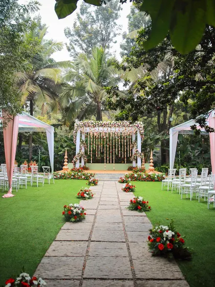A classic wedding ceremony setup with a floral mandap and flower petals lining the aisle. The tall palm trees in the background add to the tropical, garden feel.
