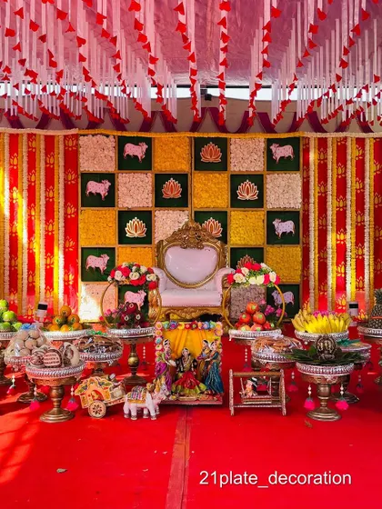 A festive outdoor baby shower setup with a red, yellow, and green theme. The decor includes a canopy, a checkered backdrop with cow motifs, and a full display of traditional items.