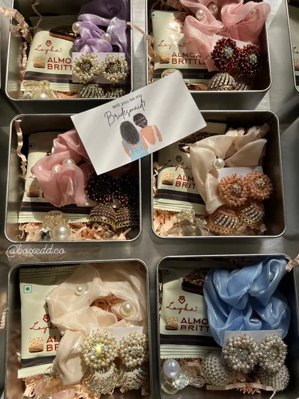 These compact tin boxes are a popular choice for bridesmaid favors. Each one is filled with a curated selection of accessories like earrings and scrunchies, along with a sweet treat and a "Will you be my Bridesmaid?" card.
