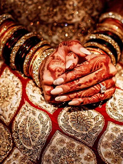 A detailed photograph of a bride's hands, showcasing her elaborate bangles and the rich embroidery of her lehenga.