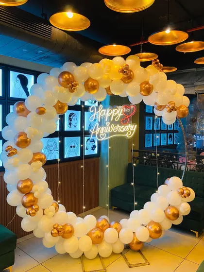 An elegant anniversary decoration for a restaurant or cafe setting. This full balloon ring in white and chrome gold is accented with fairy lights and a "Happy Anniversary" neon sign, creating a beautiful and celebratory focal point.