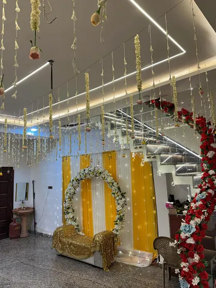 A view of the wedding house interior, showing the Haldi setup in one corner and a red floral photo wall in another, with the ceiling decorated with hanging flowers.