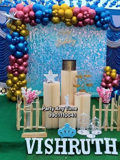 A vibrant first birthday setup with a blue shimmer wall and a colorful balloon arch in blue, yellow, and pink.