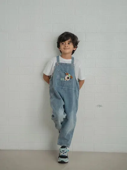 A simple and classic portrait of a young boy in denim overalls leaning against a white brick wall.