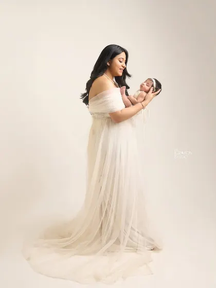 A full-length portrait of a mother in a flowing white gown, holding her newborn. It showcases the elegance and grace of new motherhood.