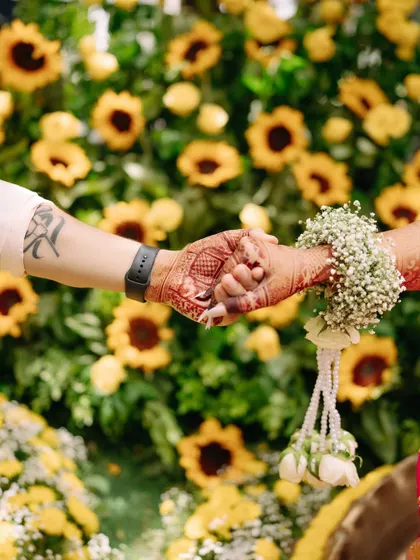 Holding hands against a backdrop of sunflowers, a beautiful detail shot from a vibrant Haldi ceremony.