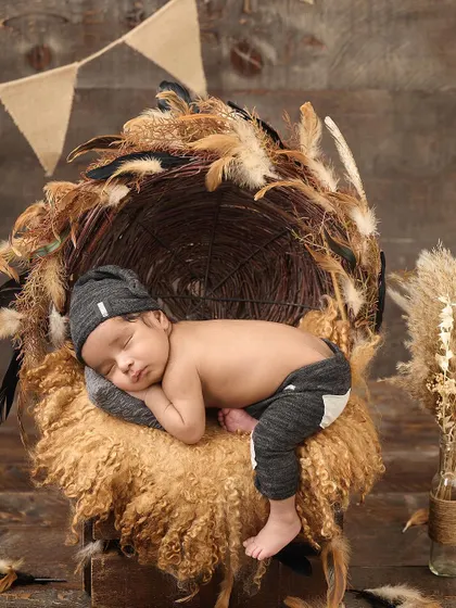 A newborn sleeps on a fluffy surface in a nest-like basket decorated with pampas grass and feathers, perfectly capturing a boho-rustic aesthetic.